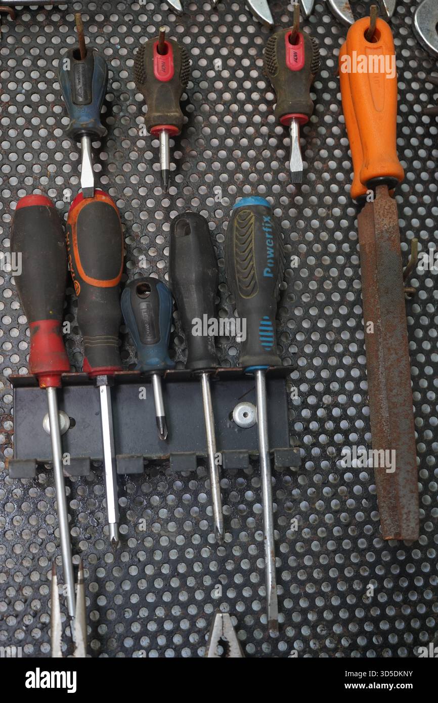 Une photo verticale de divers outils à main, y compris des tournevis et une lime avec une lame rouillée, soigneusement organisée sur un panneau perforé sombre et perforé d'atelier. Banque D'Images