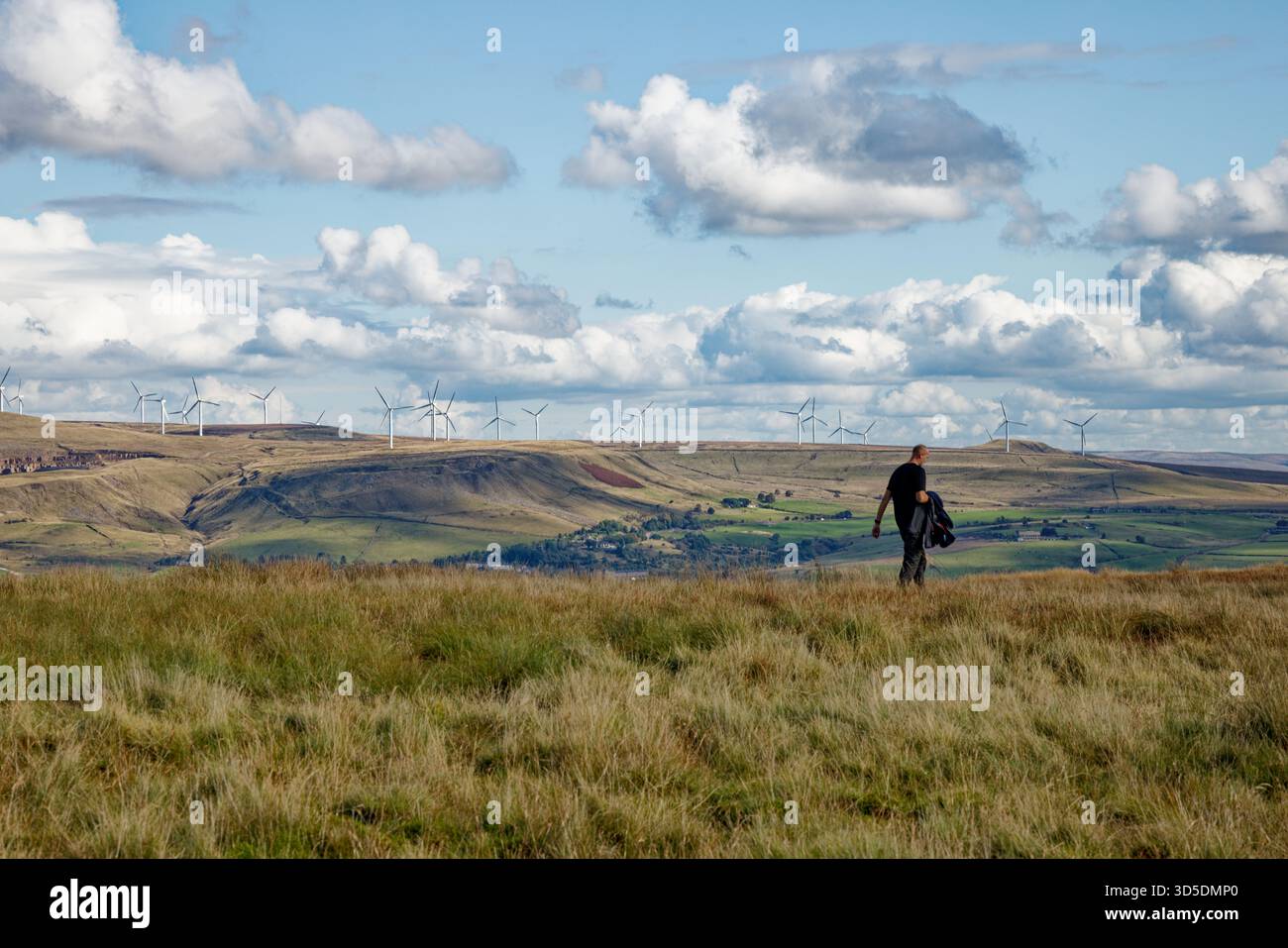 Un homme marche à travers un paysage de landes par une chaude journée avec un manteau à la main et un parc éolien sur la crête d'une colline lointaine, West Pennines, Lancashire. Banque D'Images