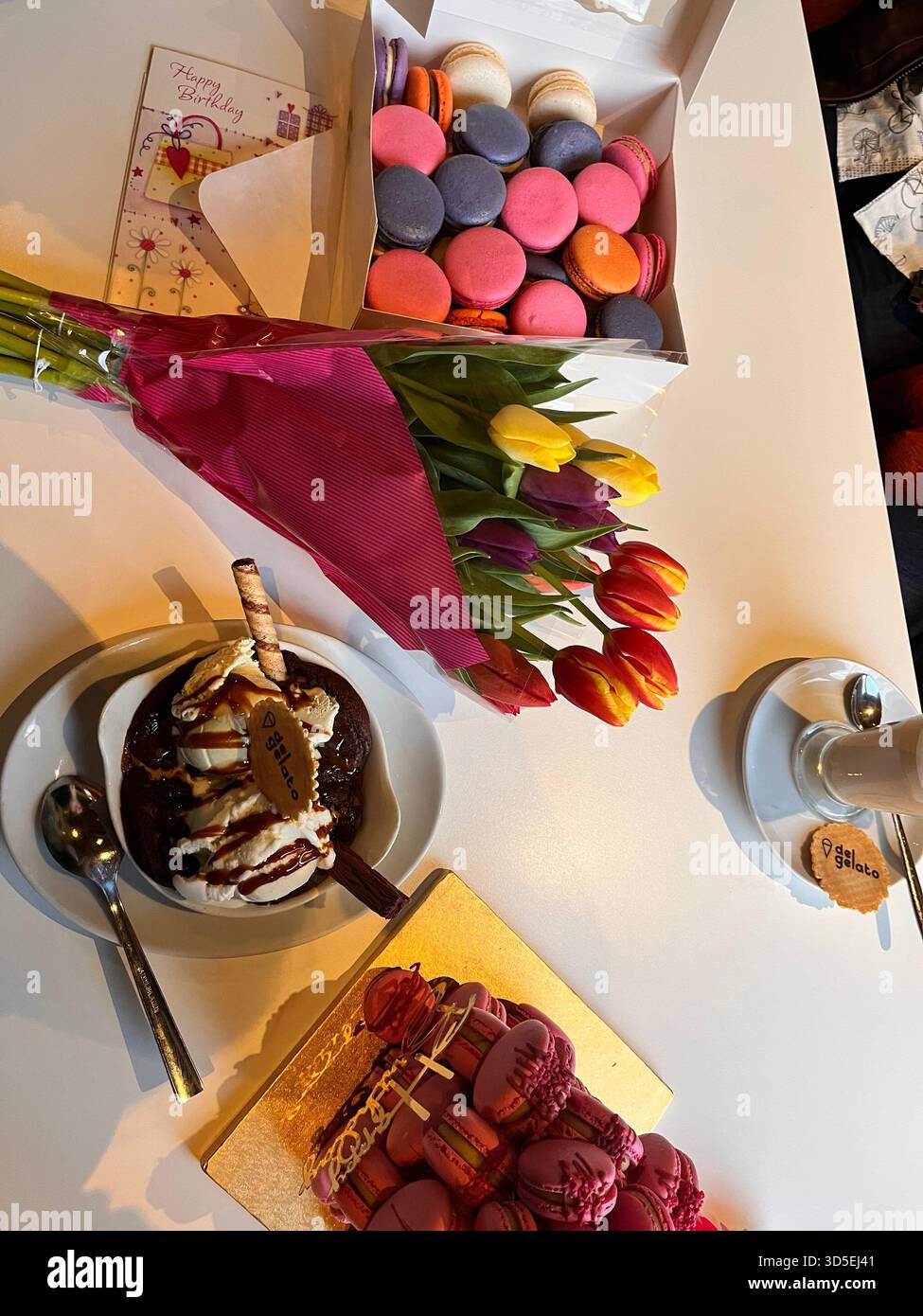 Une boîte de macarons colorés associés à un bouquet de tulipes repose sur une table lumineuse avec des desserts et du café. Banque D'Images