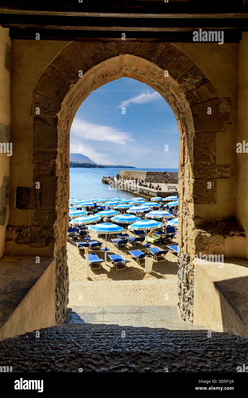 Cefalu, Italie -19 septembre 2025 : vue sur la plage de Cefalu avec des parasols bleus et une jetée en bord de mer encadrée par une ancienne arche en pierre par une journée ensoleillée à Sicil Banque D'Images