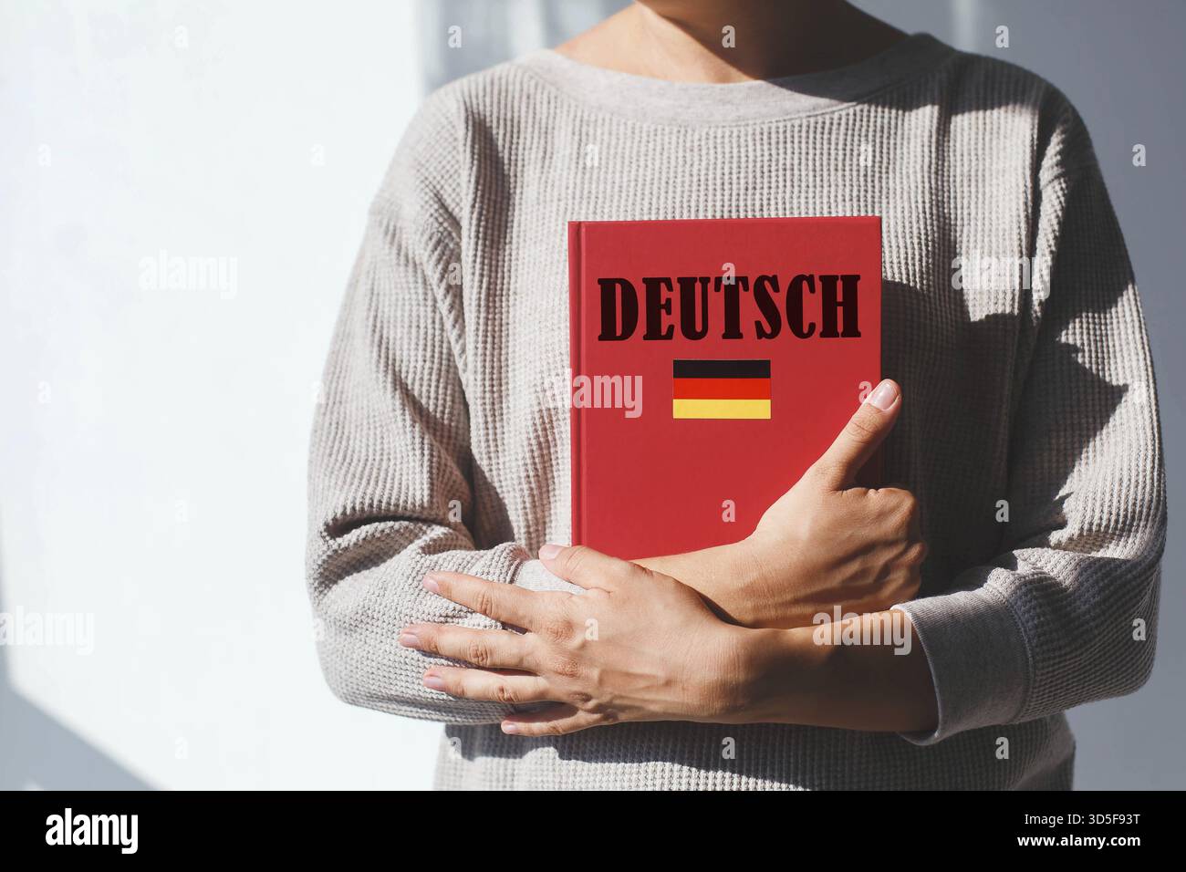 Jeune femme en pull beige tient un manuel de langue allemande rouge avec inscription allemande et drapeau allemand, concept d'apprentissage des langues étrangères. son visage i Banque D'Images