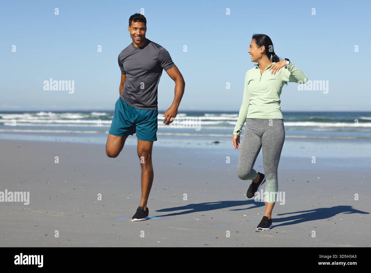 Couple s'exerçant sur la plage, étirant les jambes et souriant dans la lumière du soleil du matin Banque D'Images