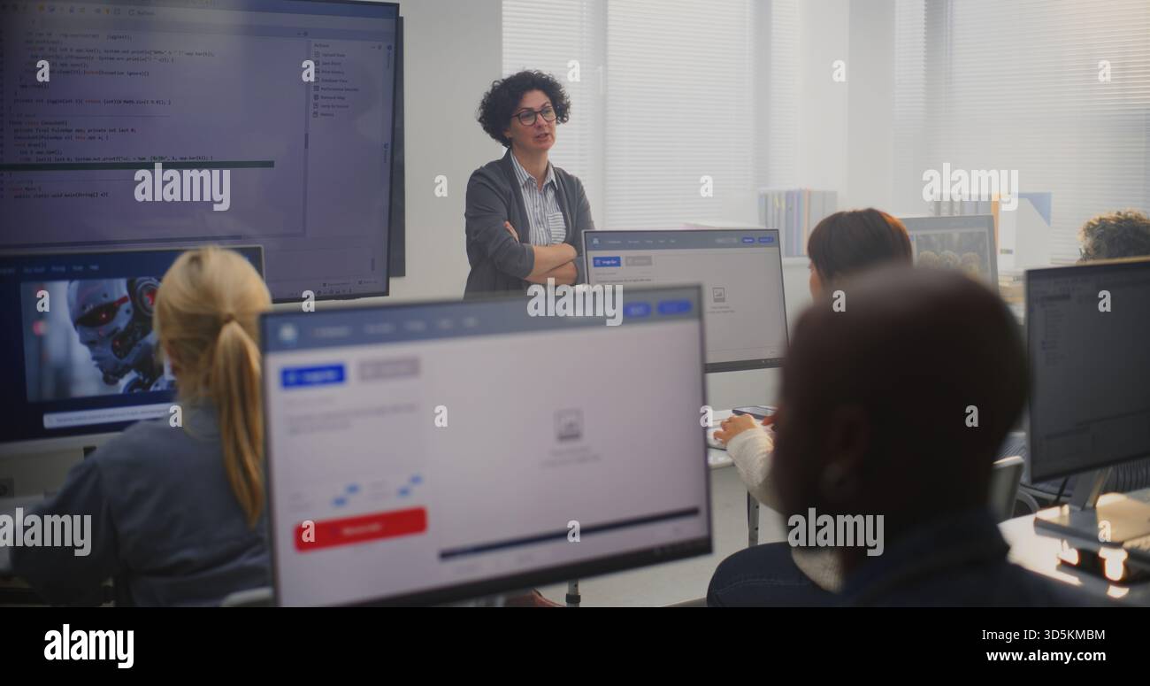 Enseignante se tient à côté du grand écran affichant le code de programmation, expliquant les concepts de modélisation aux élèves adultes. Concept éducation numérique, compétences en codage, environnement d'apprentissage professionnel moderne. Banque D'Images