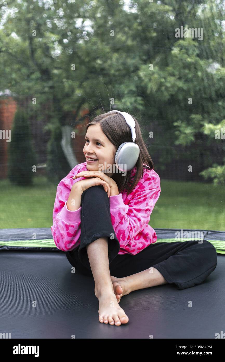 Souriant fille de 8 à 9 ans en sweat-shirt imprimé léopard rose et écouteurs assis pieds nus sur un trampoline, concept de relaxation de l'enfance, thème o Banque D'Images