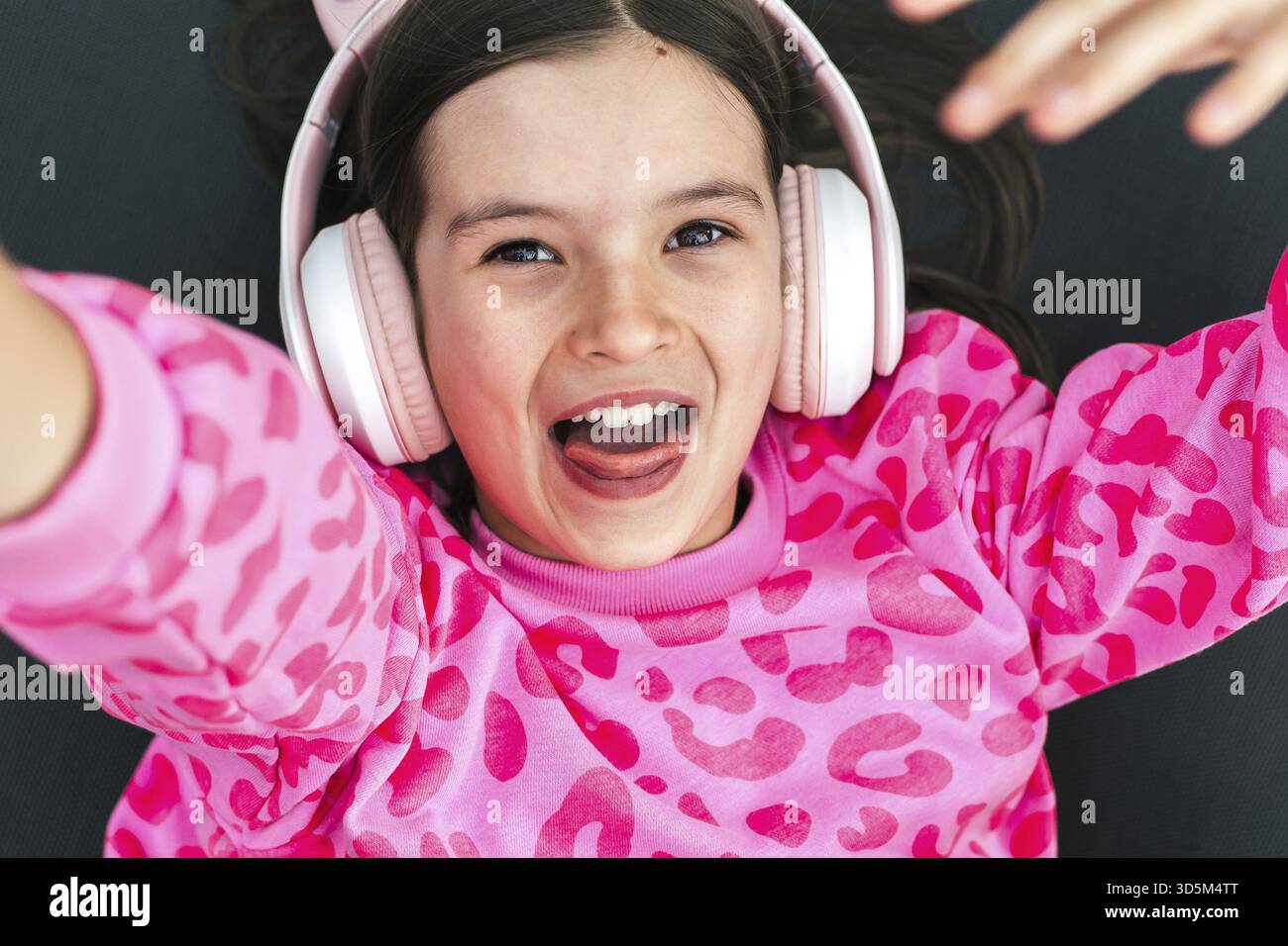 Joyeuse fille de 8-9 ans en sweat-shirt imprimé léopard rose et écouteurs couchés et souriants joyeusement, concept d'énergie de l'enfance, thème du plaisir, Banque D'Images