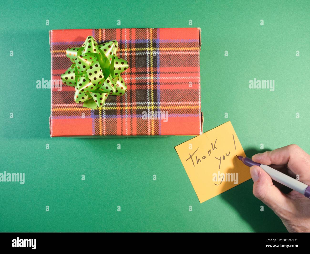 Boîte cadeau de remerciement avec note manuscrite - vue de dessus d'une boîte cadeau festive à carreaux avec un noeud vert et une main écrivant un « Merci ! » note sur un jaune Banque D'Images