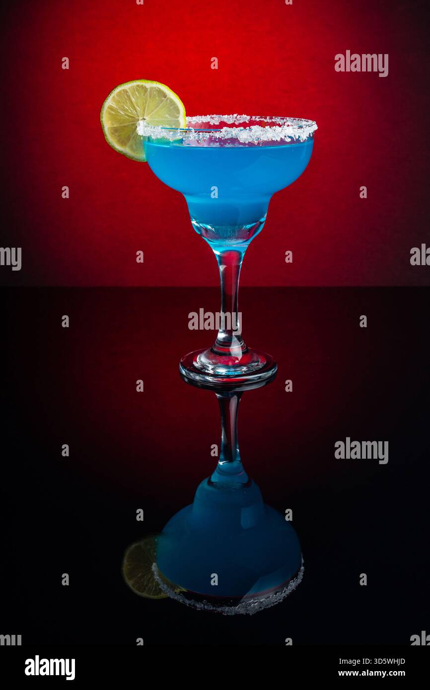 Un cocktail bleu dans un verre à margarita avec bordure salée et tranche de citron vert, reflété sur la surface noire. Banque D'Images