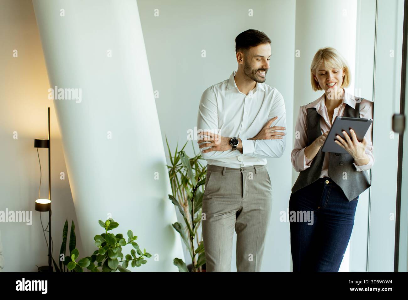 Deux collègues profitent d'un moment de lumière en examinant les détails du projet sur une tablette tout en se tenant debout près de grandes fenêtres. Leurs sourires montrent un travail positif envi Banque D'Images