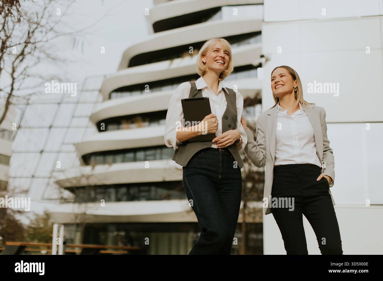 Deux femmes en tenue d'affaires sourient et discutent en marchant à l'extérieur d'un bâtiment contemporain. Ils portent une tablette et dégagent une énergie positive, mettant en valeur Banque D'Images