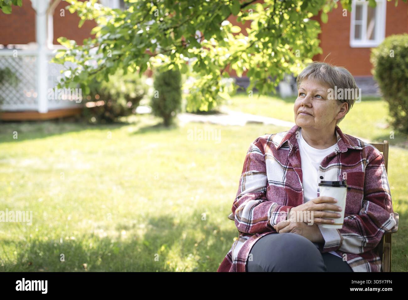 Femme âgée en chemise à carreaux rouge et t-shirt blanc assis dans le jardin avec une tasse à café sur une journée ensoleillée, regardant réfléchi, concept de réflexion, thème Banque D'Images