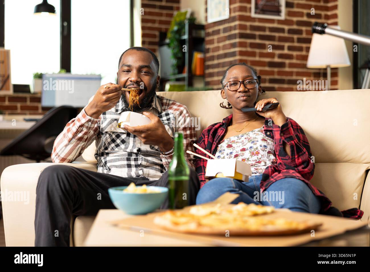 Homme barbu appréciant les nouilles comme petite amie tient à distance, à la fois la liaison sur l'émission de télévision dans l'appartement de mur de briques. Jeune couple noir ayant une ambiance détendue week-end sur le canapé confortable du salon. Banque D'Images