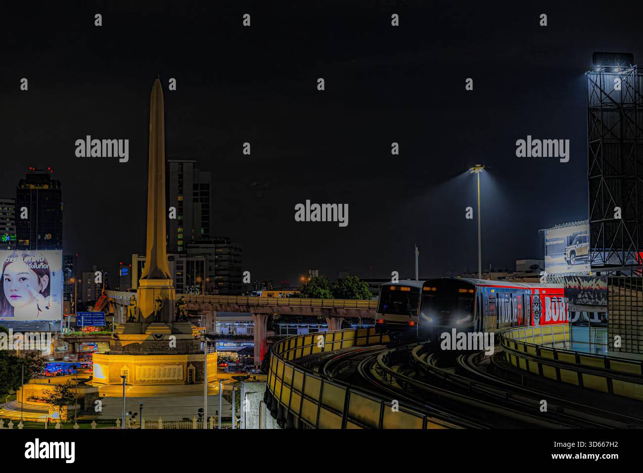Vue nocturne du Monument de la victoire à Bangkok avec un Skytrain BTS passant au-dessus de vous, capturant l'atmosphère urbaine vibrante et le mouvement constant autour Banque D'Images