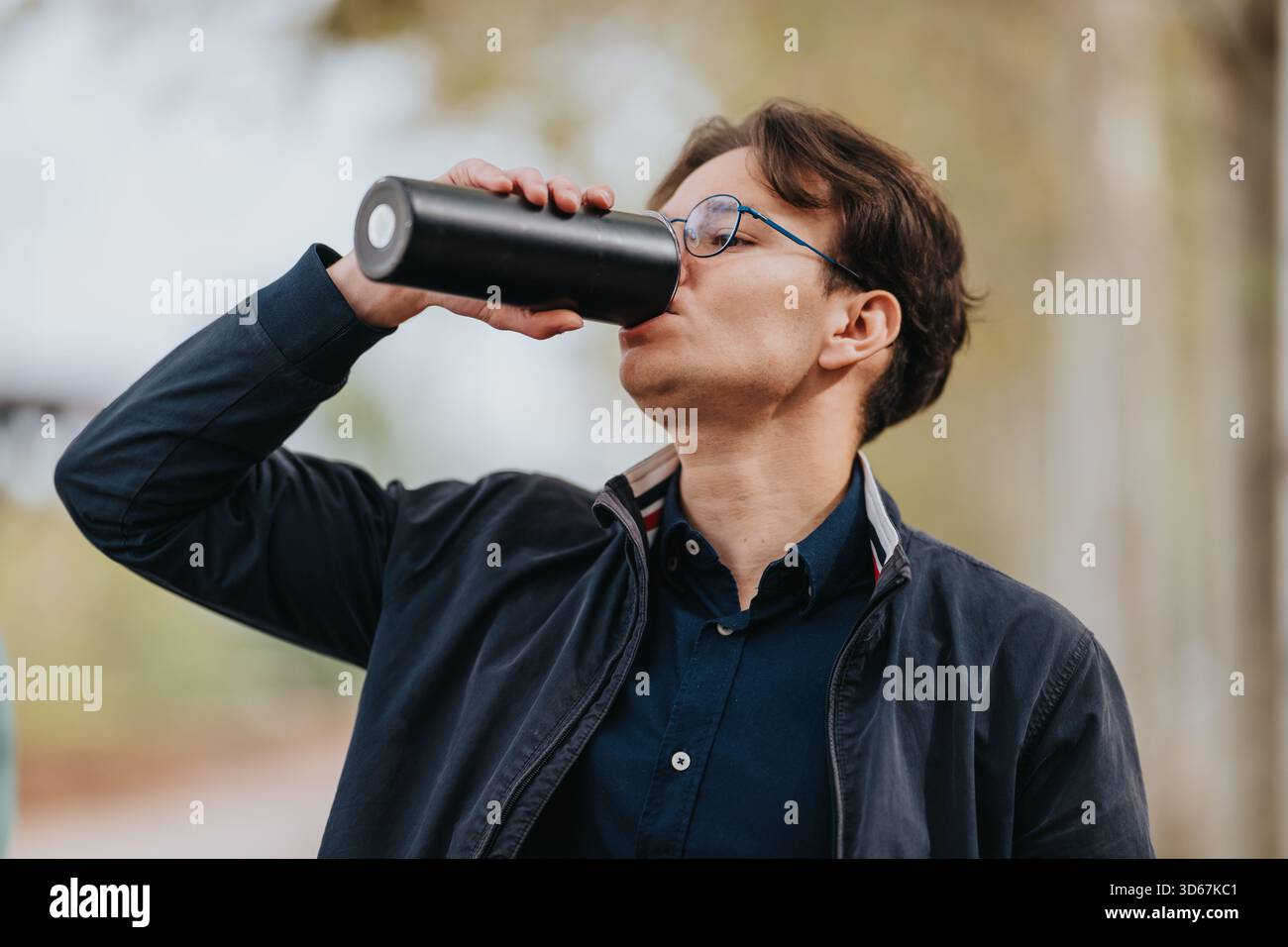 L'homme boit de l'eau d'une bouteille à l'extérieur dans une veste décontractée et des verres pendant une promenade dans le parc Banque D'Images