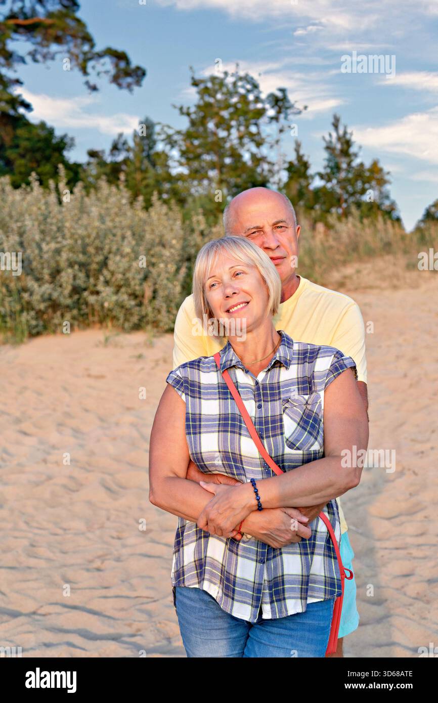Couple plus âgé souriant embrassant sur la plage de sable au coucher du soleil, lumière chaude heure dorée et ambiance estivale détendue Banque D'Images