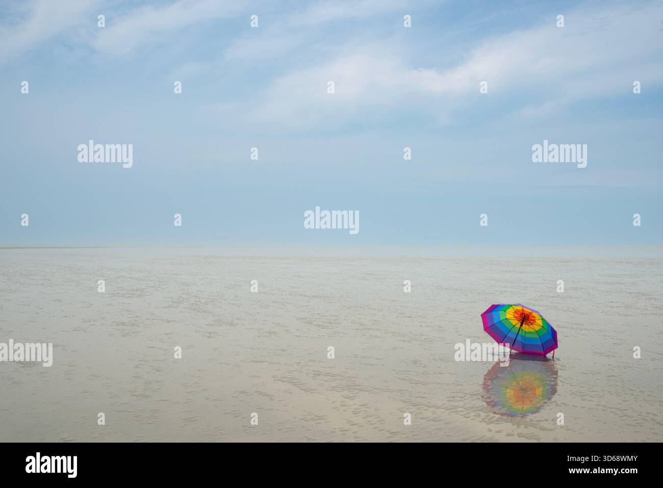 Un parapluie solitaire et éclatant de couleur arc-en-ciel se trouve sur une vase humide et sablonneuse sous un ciel doux et nuageux, créant un reflet. Banque D'Images