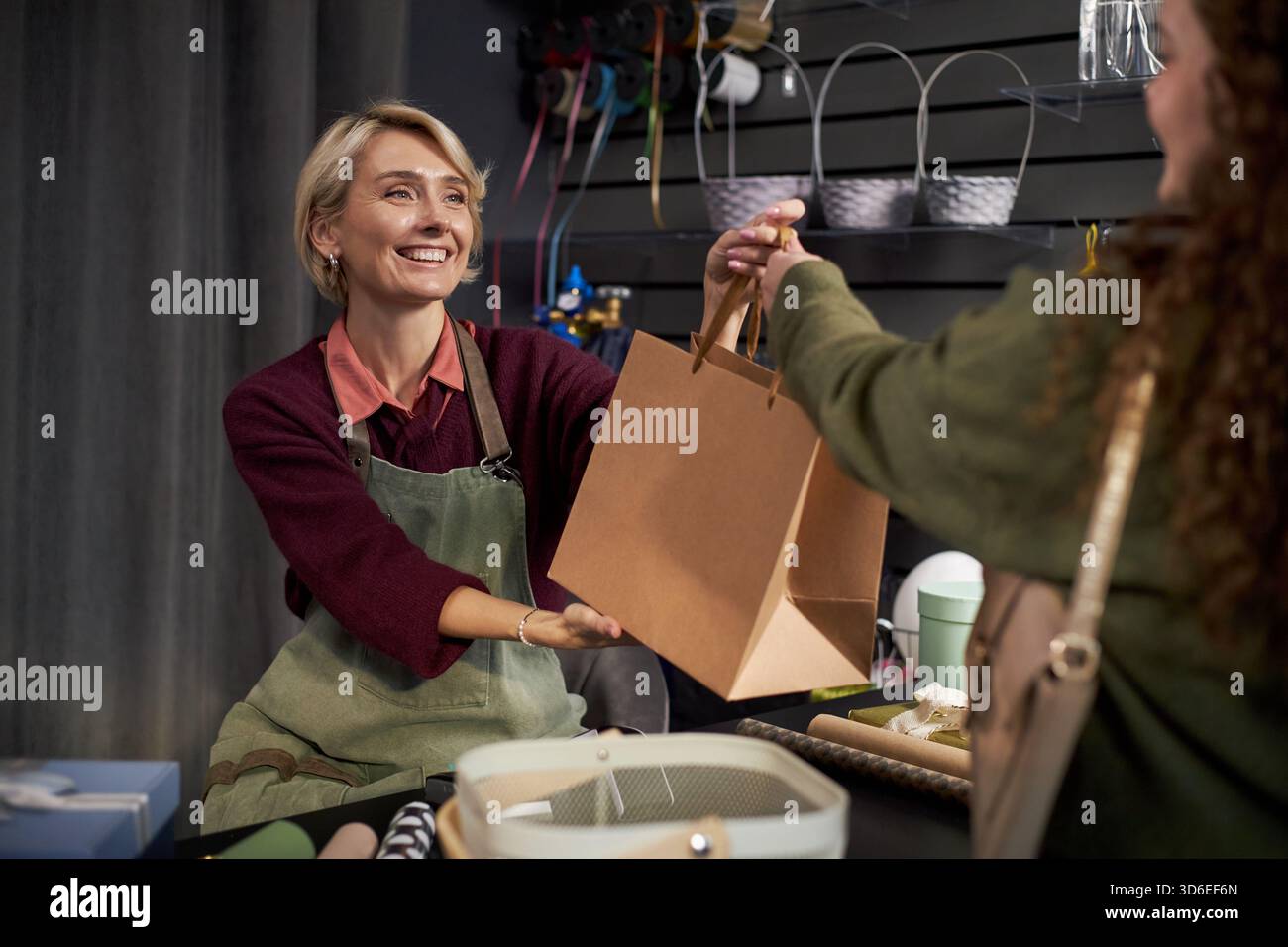Femme d'âge moyen caucasien souriant tout en remettant un sac à provisions en papier à la jeune femme adulte dans un magasin de fleurs, comptoir affichant des fournitures florales et des matériaux d'emballage en arrière-plan Banque D'Images