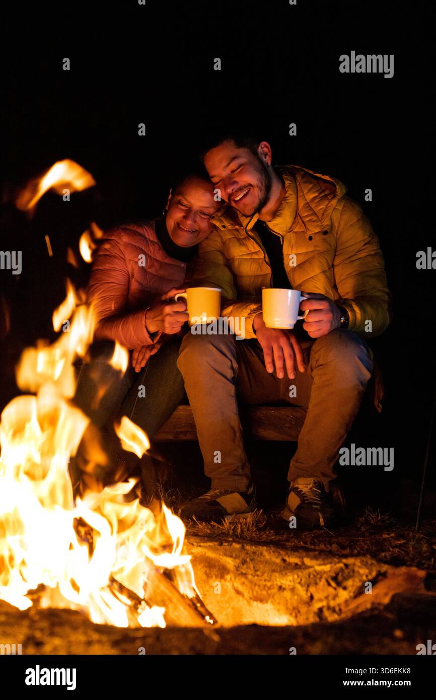 Une scène confortable par le feu de camp la nuit, soulignant la chaleur. Deux personnes apprécient les boissons chaudes dans les feux de lueur, portant des vestes contre le froid dans le fo Banque D'Images