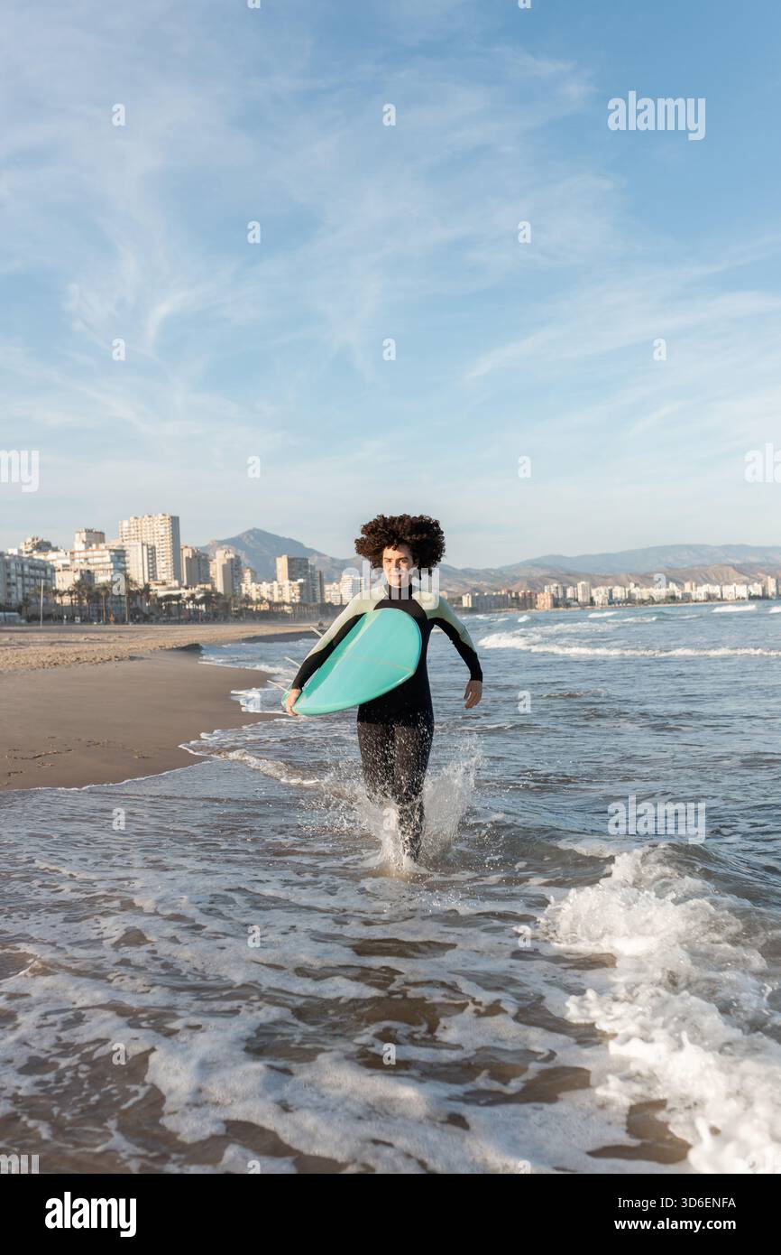 Jeune femme de surf attentionnés en combinaison avec planche de surf qui s'étourne sur la mer lavée par la mer Banque D'Images