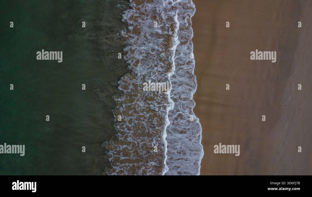 Des vagues blanches mousseuses se brisent entre le sable sombre et l'eau profonde de l'océan vert émeraude Banque D'Images
