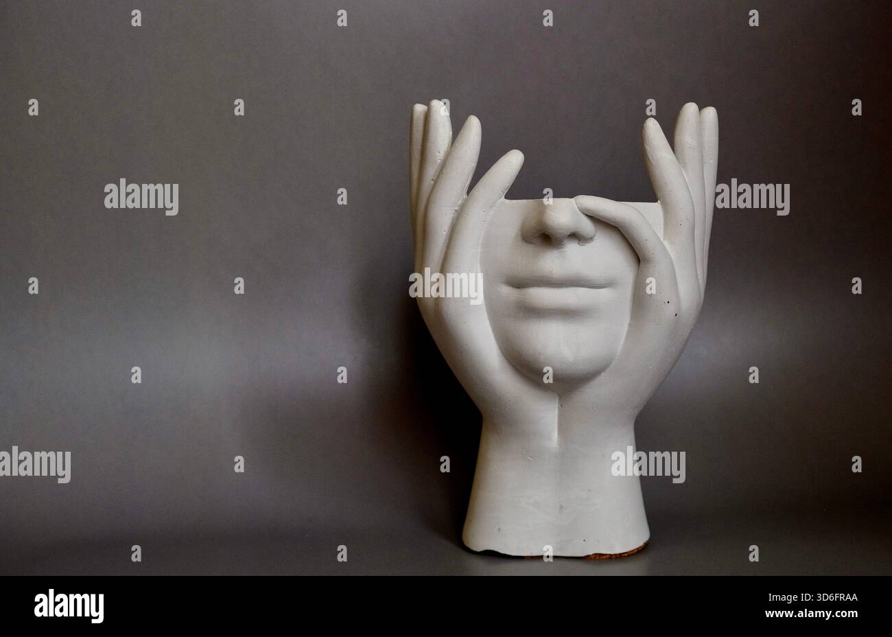 Une sculpture de visage en plâtre encadrée par deux mains, positionnée sur un fond gris neutre. Nature morte artistique minimaliste avec symbolisme émotionnel. Banque D'Images