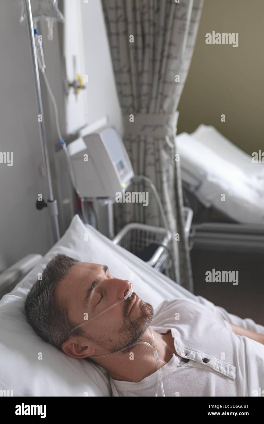 Homme d'âge moyen couché dans un lit d'hôpital, portant un henley blanc, recevant de l'oxygène via une canule nasale. Patient, service, rideau, moniteur, perfusion, repos, Banque D'Images