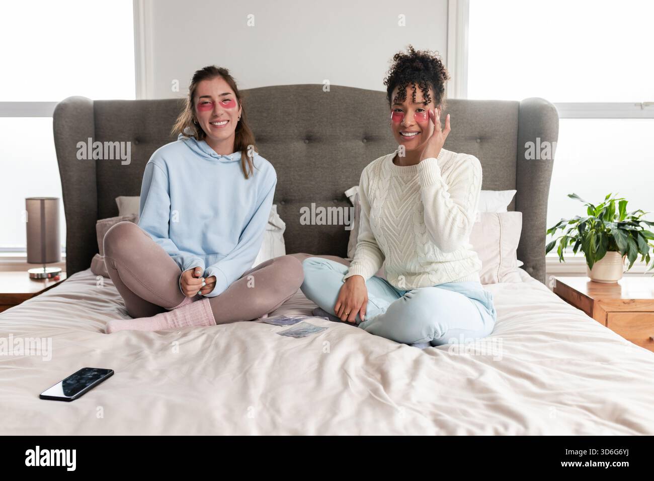 Amies féminines diverses appliquant des patchs de gel sous les yeux tout en étant assis les jambes croisées sur un lit king-size Banque D'Images