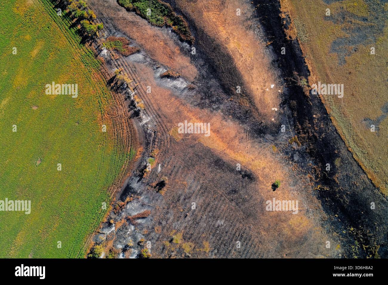 Photographie aérienne descendante capturant de graves dommages au sol. Motif abstrait montrant de la terre grillée noire avec des taches orange et des traces de pneus distinctes, contrastant fortement avec les terres agricoles vertes vibrantes. Banque D'Images