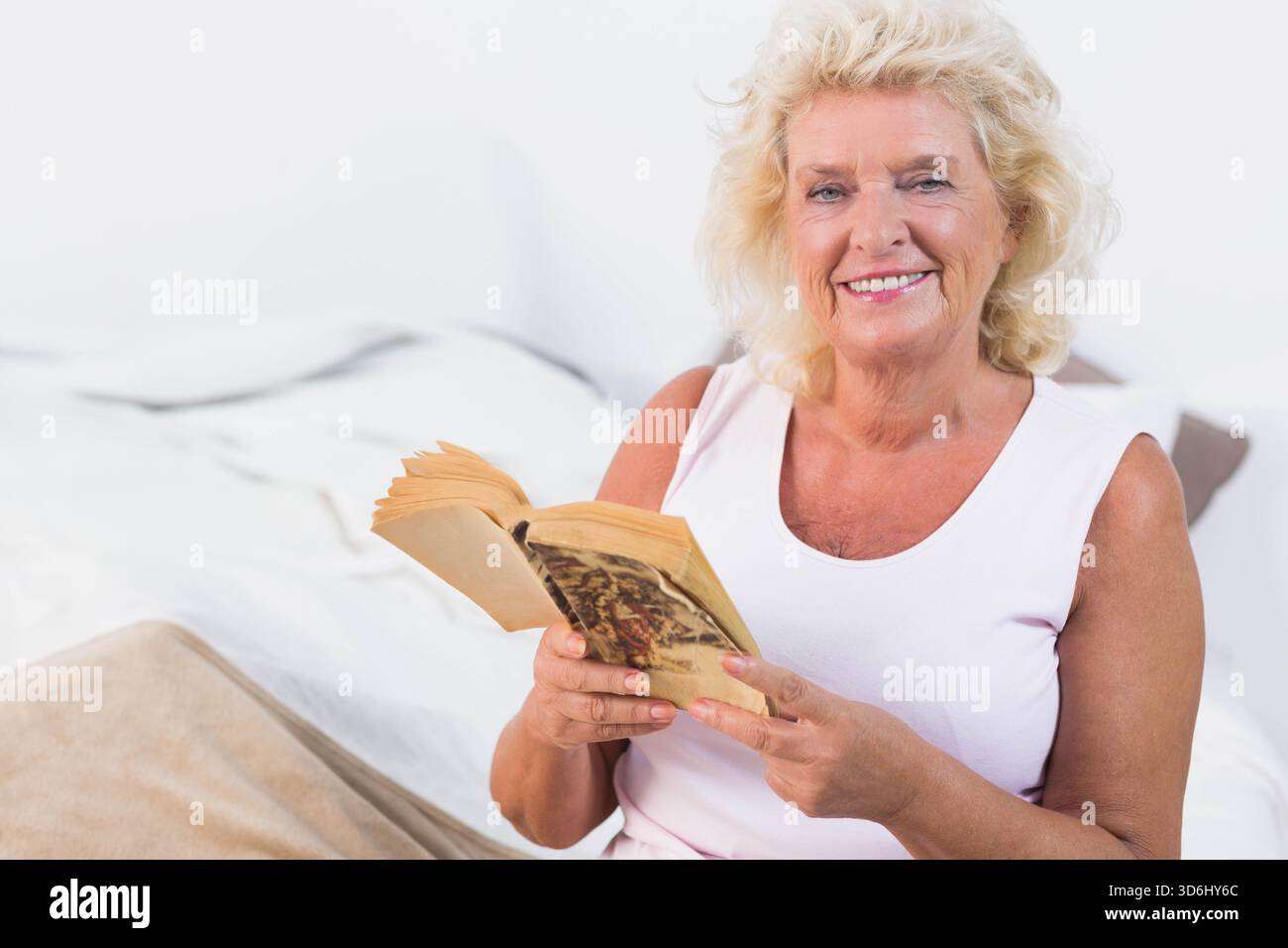Femme senior assise sur le lit avec une couverture beige portant le dessus tenant le livre de poche, souriant, espace de copie. Lecture, livre, mature, confortable, calme, lumière douce, maison Banque D'Images