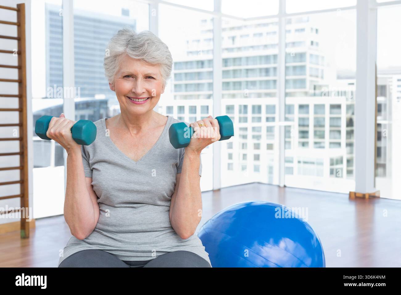 Femme senior soulevant des haltères sarcelle dans un studio de fitness, portant un T-shirt gris clair et des leggings foncés. Urbain, Highrise, skyline, lumière du jour, fenêtres, mini Banque D'Images