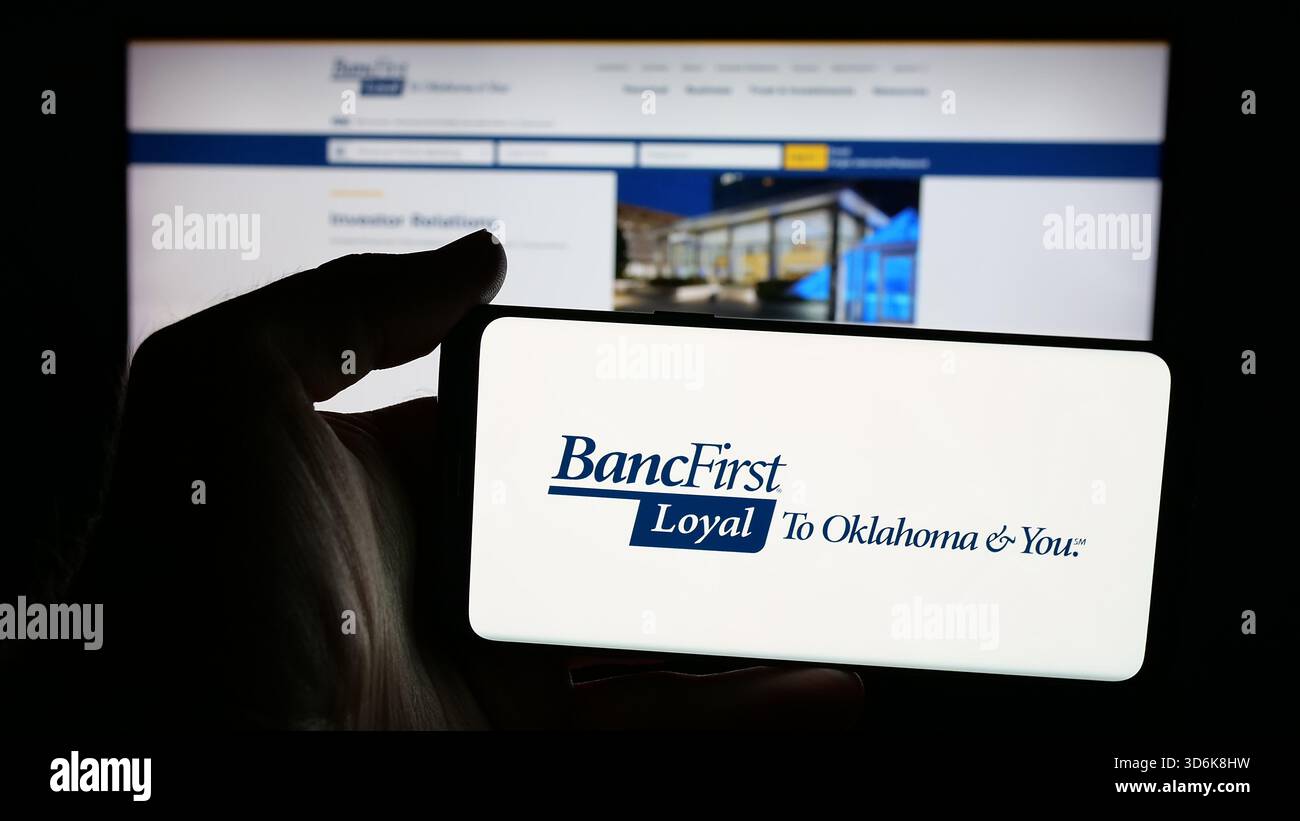 Stuttgart, Allemagne - 11-12-2025 : personne tenant un téléphone portable avec le logo de la société financière américaine BancFirst Corporation sur l'écran en face de la page Web. Banque D'Images
