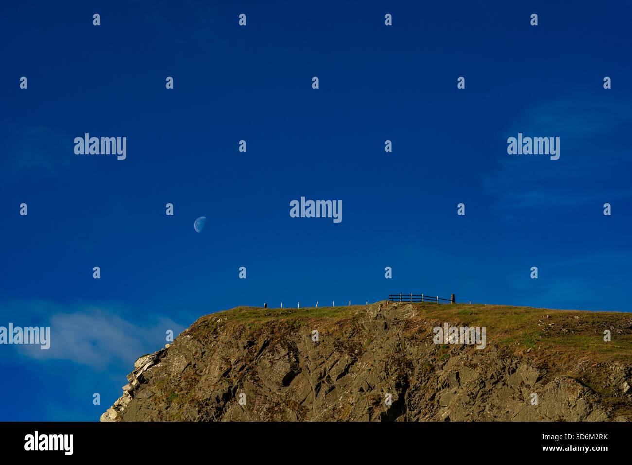 Falaise côtière surmontée d'herbe illuminée par une lumière à angle faible avec la lune suspendue dans un ciel bleu clair et profond Banque D'Images