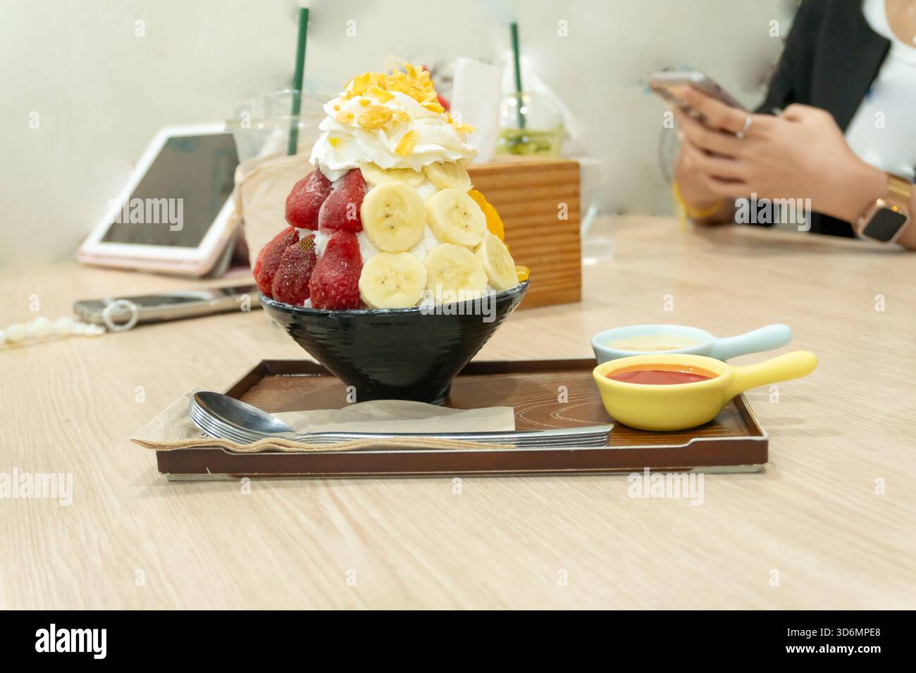 Un ensemble de Bingsu coréen dans un petit bol noir avec plusieurs fruits garniture et sauce rouge avec lait condensé sucré dans des mini bols en plastique dans un plateau brun Banque D'Images
