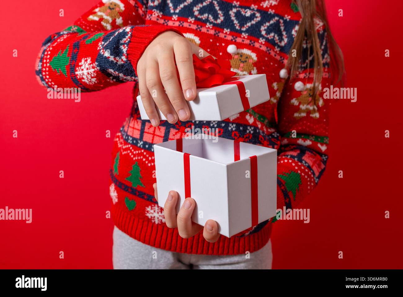 Enfant soulevant le couvercle d'une boîte cadeau blanche avec ruban rouge sur un fond rouge vif, parfait pour les promotions festives et les actifs créatifs des fêtes Banque D'Images