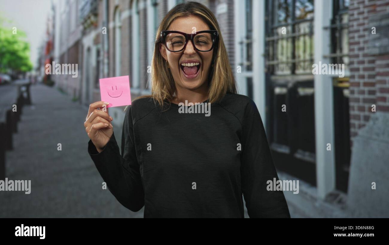 Femme tient une note collante rose avec le sourire dessiné, souriant avec la main visible sur une rue de la ville par bâtiment de briques et devanture de magasin ; positivité de joie. Banque D'Images