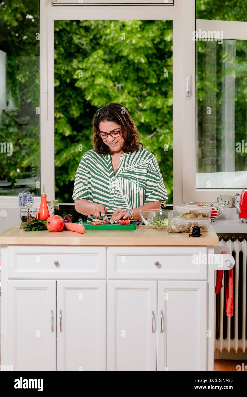 Midlife femme sourit en hachant des légumes au comptoir de la cuisine Banque D'Images