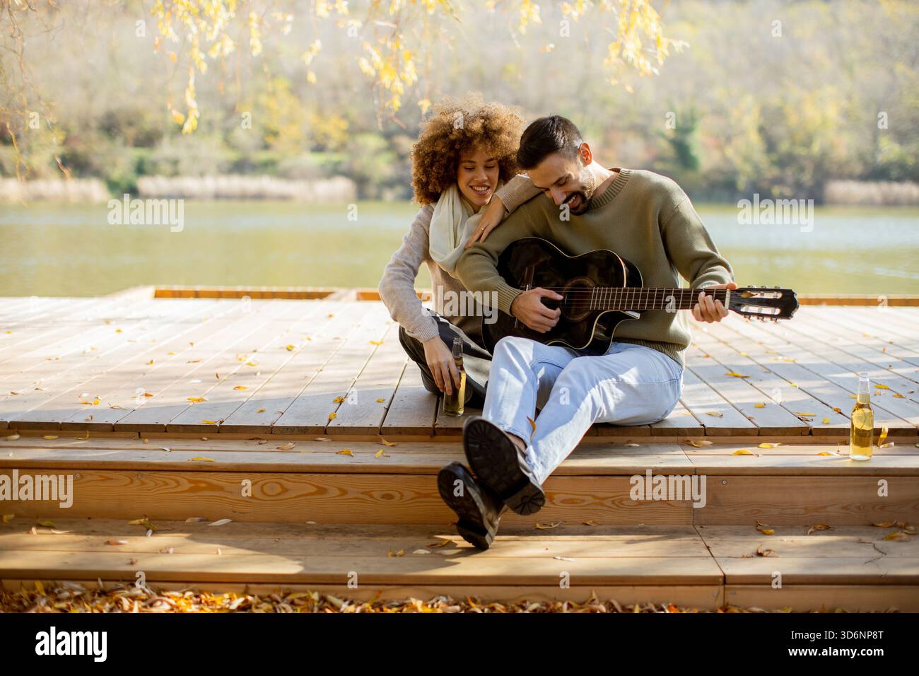 Un couple joyeux est assis sur une terrasse en bois au bord d'un lac serein, partageant des sourires tandis que l'un caresse une guitare et l'autre écoute, entouré de feuilles d'automne. Banque D'Images