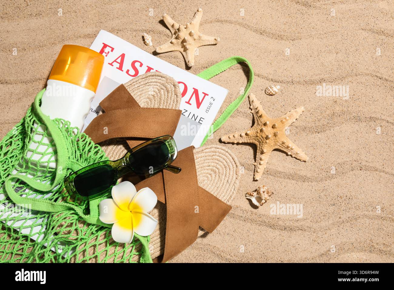 Sac à ficelle avec bouteille de crème solaire, magazine et accessoires de plage sur sable comme fond, gros plan Banque D'Images