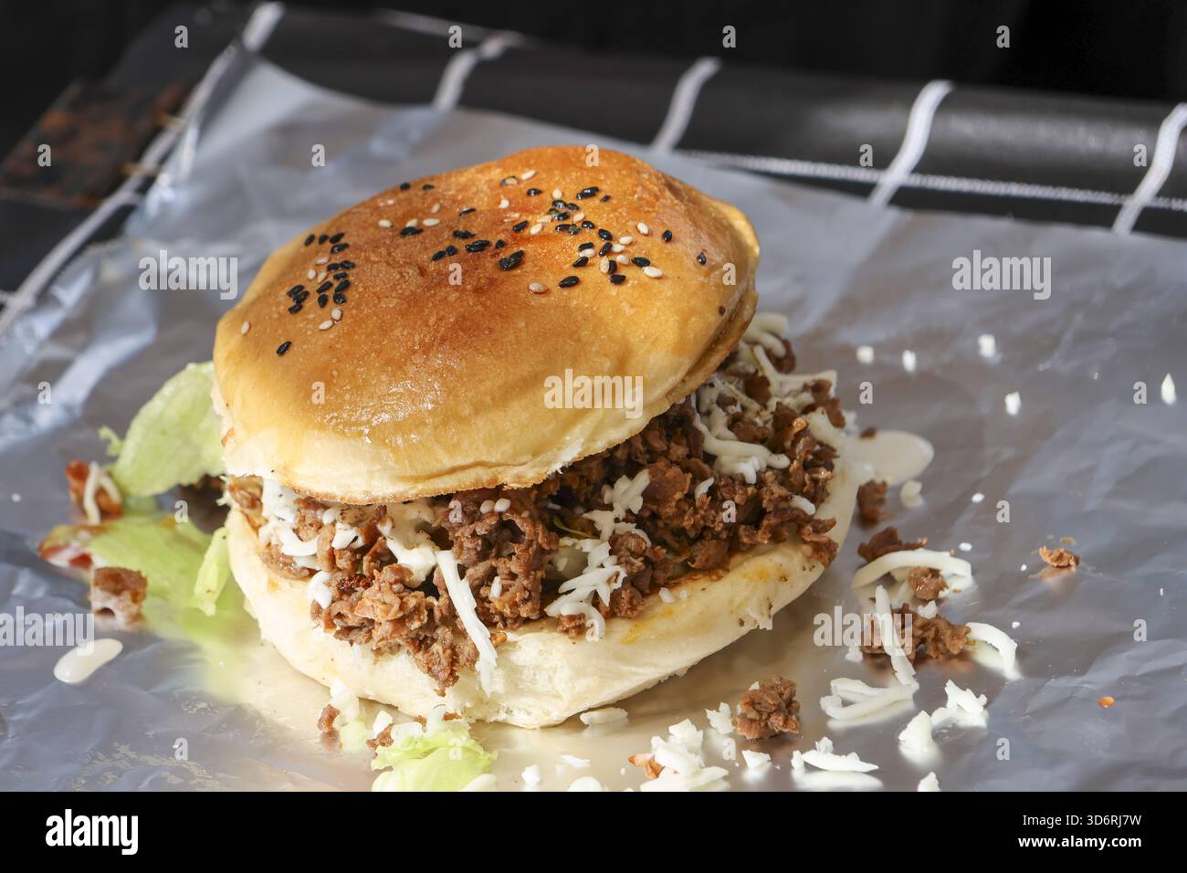 Sandwich hamburger de bœuf haché en désordre sur pain aux graines de sésame avec sauce crémeuse. Restauration rapide appétissante pour un déjeuner ou un dîner copieux, servie sur du papier aluminium à emporter Banque D'Images
