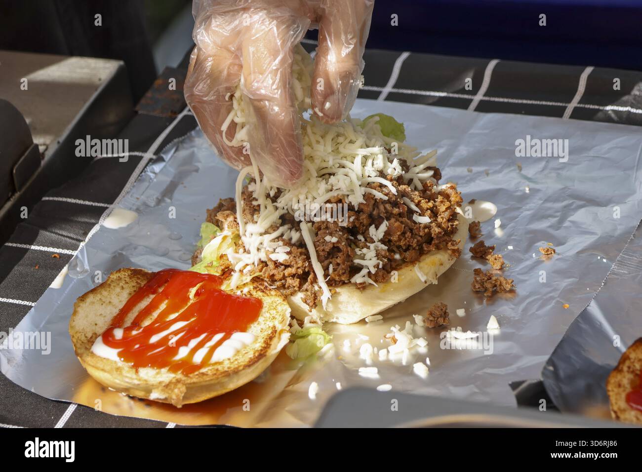 Chef préparant un délicieux sandwich Street food avec viande hachée, fromage râpé et sauce. Préparation des aliments en gros plan montre la main dans l'assemblage du gant Banque D'Images