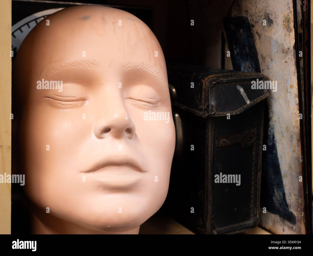 La tête de mannequin avec les yeux fermés repose à côté d'un étui en cuir usé sur une étagère, légèrement éclairée pour souligner l'expression calme et la texture de la surface. Banque D'Images