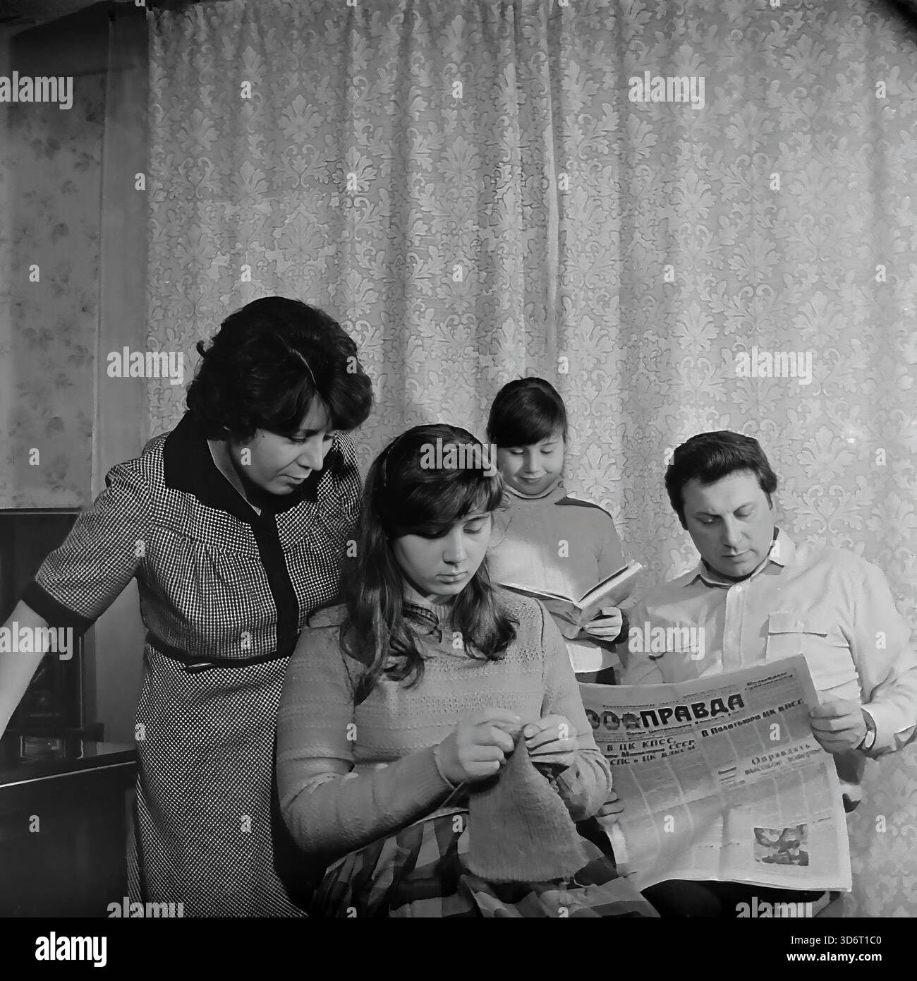 Slaviansk, région de Donetsk, RSS d'Ukraine, Union soviétique. Février 1986. La famille du député Kolesnik passant une soirée à la maison. Le père lit le journal de la Pravda tandis que la mère regarde leur fille aînée tricoter avec de la laine. La jeune fille se tient derrière la lecture d'un livre. Ils portent des vêtements décontractés dans une pièce avec des rideaux à motifs. Cette photographie d'archives en noir et blanc capture une scène de loisirs domestiques traditionnels dans la paisible région du Donbass bien avant la guerre. Banque D'Images
