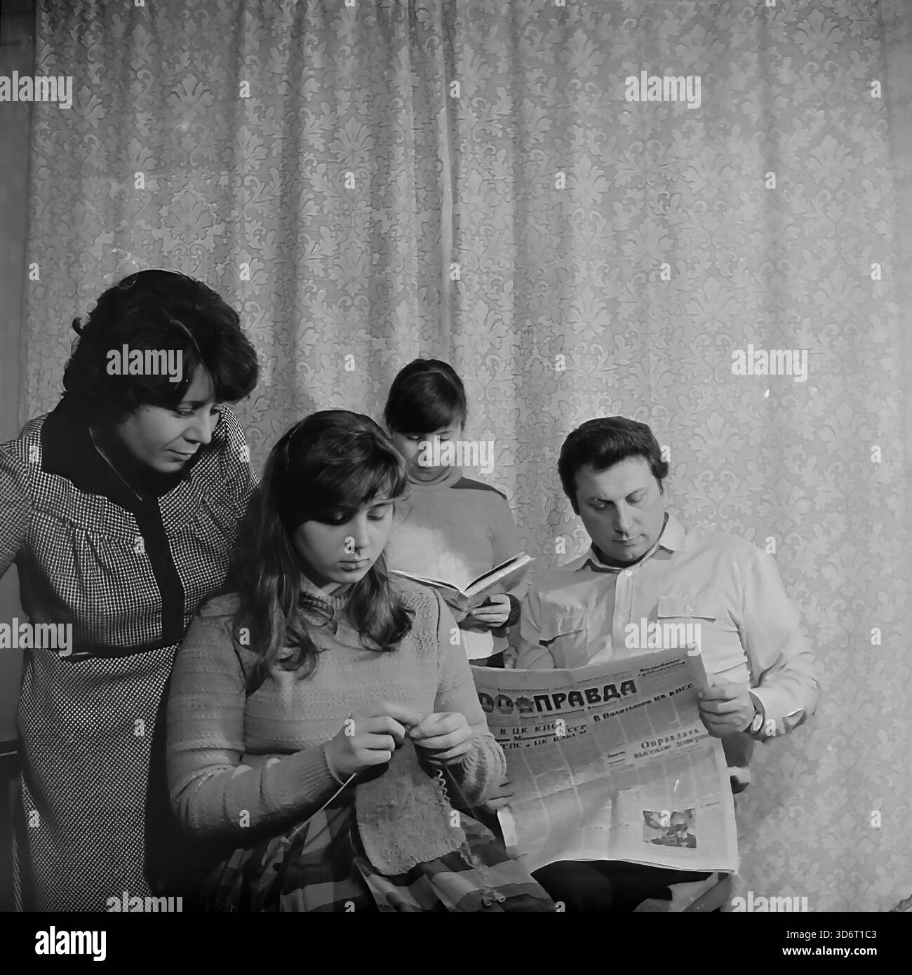 Slaviansk, région de Donetsk, RSS d'Ukraine, Union soviétique. Février 1986. La famille du député Kolesnik passant une soirée à la maison. Le père lit le journal de la Pravda tandis que la mère regarde leur fille aînée tricoter avec de la laine. La jeune fille se tient derrière la lecture d'un livre. Ils portent des vêtements décontractés dans une pièce avec des rideaux à motifs. Cette photographie d'archives en noir et blanc capture une scène de loisirs domestiques traditionnels dans la paisible région du Donbass bien avant la guerre. Banque D'Images