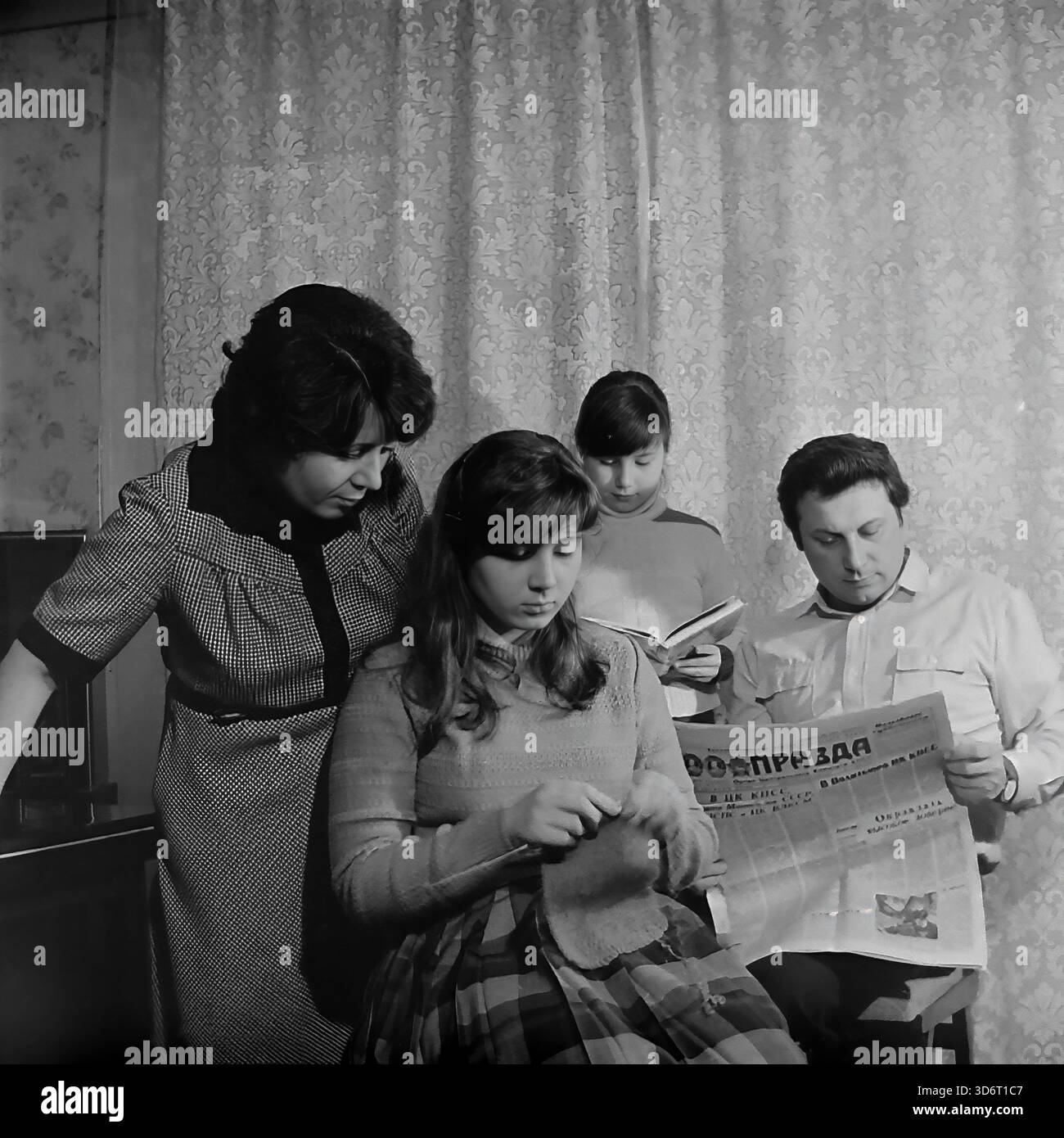Slaviansk, région de Donetsk, RSS d'Ukraine, Union soviétique. Février 1986. La famille du député Kolesnik passant une soirée à la maison. Le père lit le journal de la Pravda tandis que la mère regarde leur fille aînée tricoter avec de la laine. La jeune fille se tient derrière la lecture d'un livre. Ils portent des vêtements décontractés dans une pièce avec des rideaux à motifs. Cette photographie d'archives en noir et blanc capture une scène de loisirs domestiques traditionnels dans la paisible région du Donbass bien avant la guerre. Banque D'Images