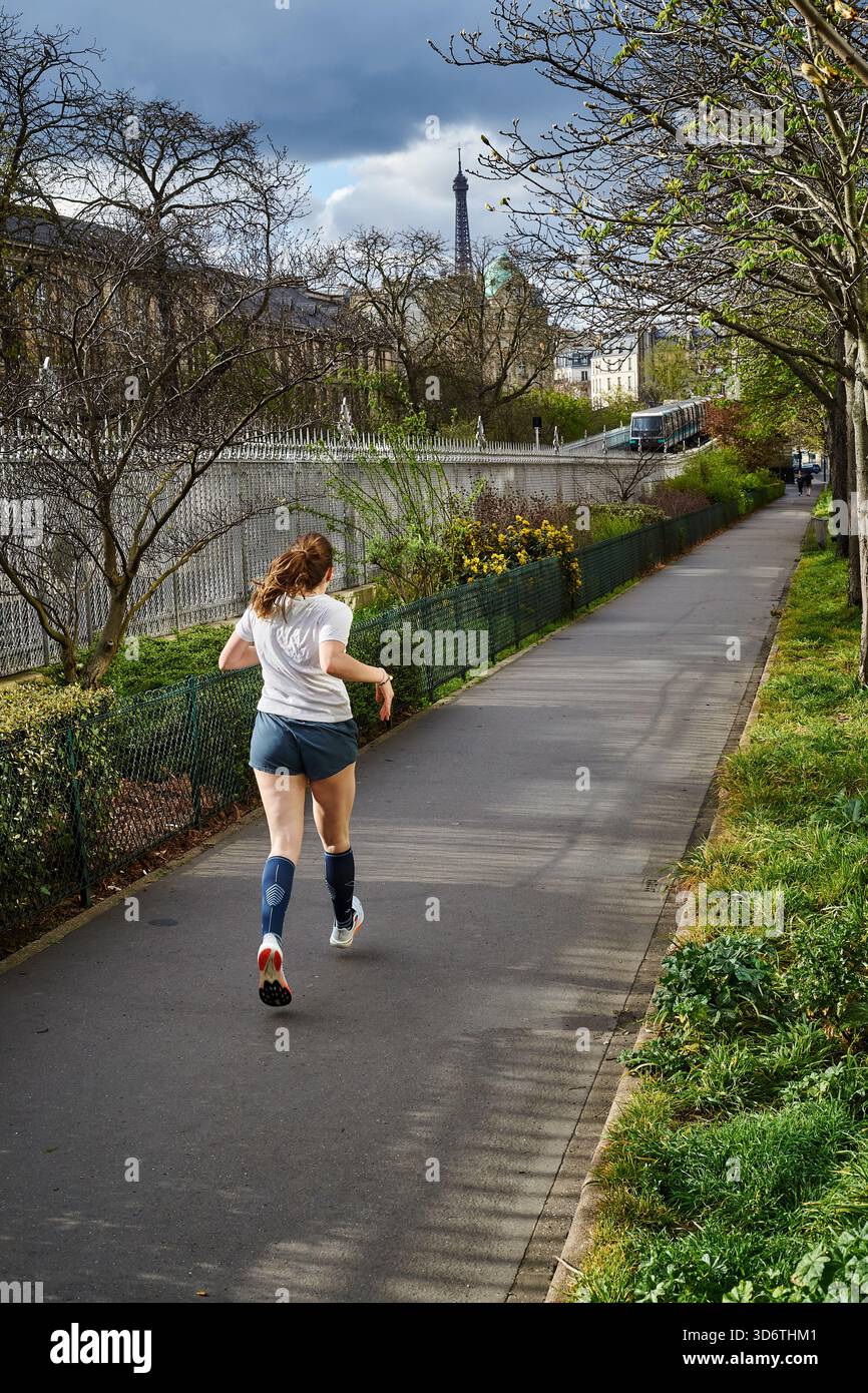 Paris, France - mars 2025 : une femme fait du jogging le long d'un sentier bordé d'arbres dans le 15e arrondissement de Paris. Le soleil brille à travers les nuages, créant un brick Banque D'Images