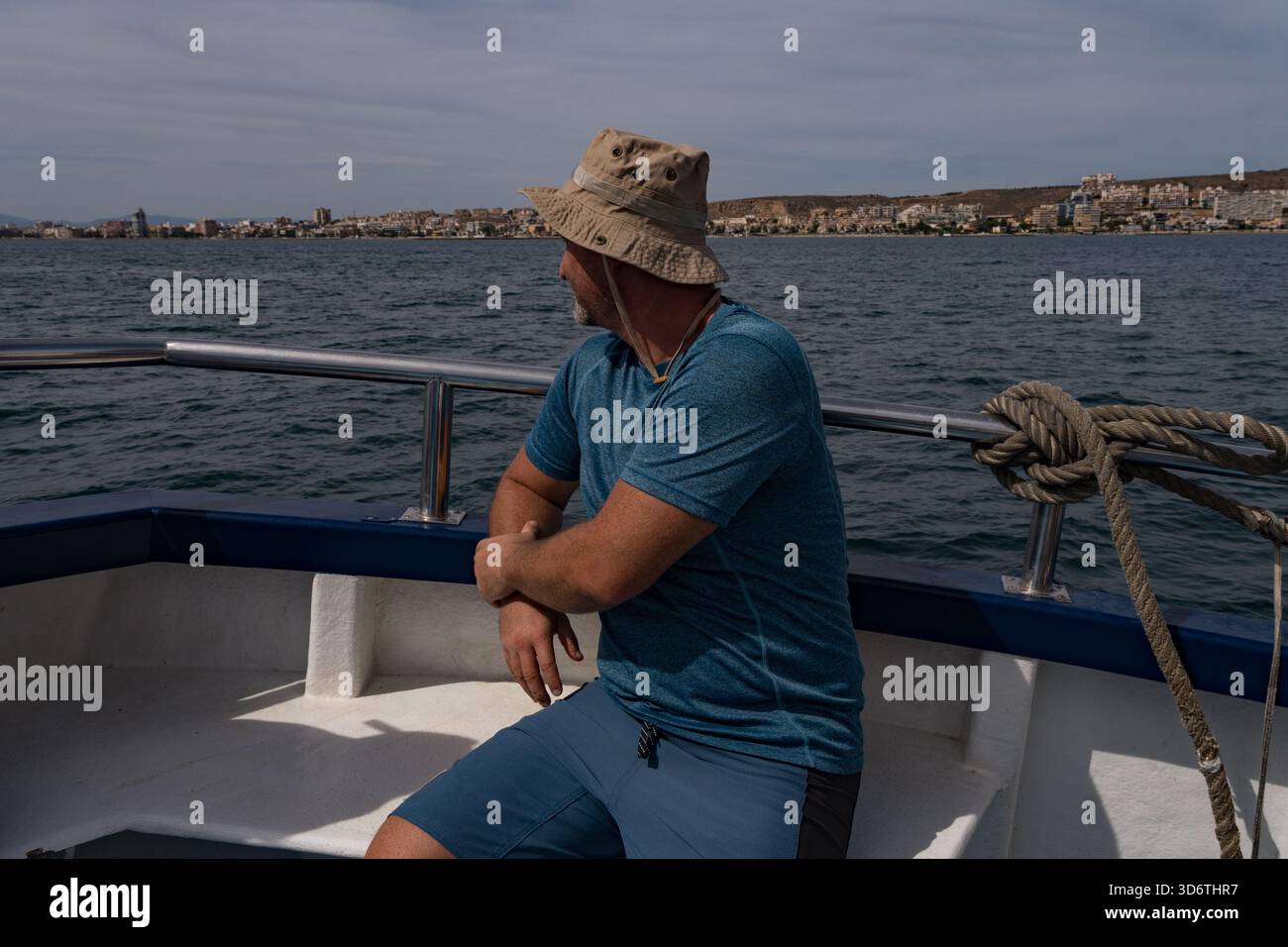 Homme détendu assis sur le bord d'un voilier, encadré par la mer bleue et gréement, profitant d'une journée ensoleillée sur l'eau. Regardant devant lui. Banque D'Images