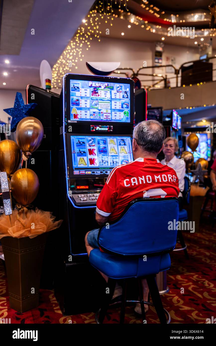 Homme assis à une machine à sous de casino entouré de lumières festives et décor de jeu, créant une atmosphère de jeu et de divertissement animée. Banque D'Images