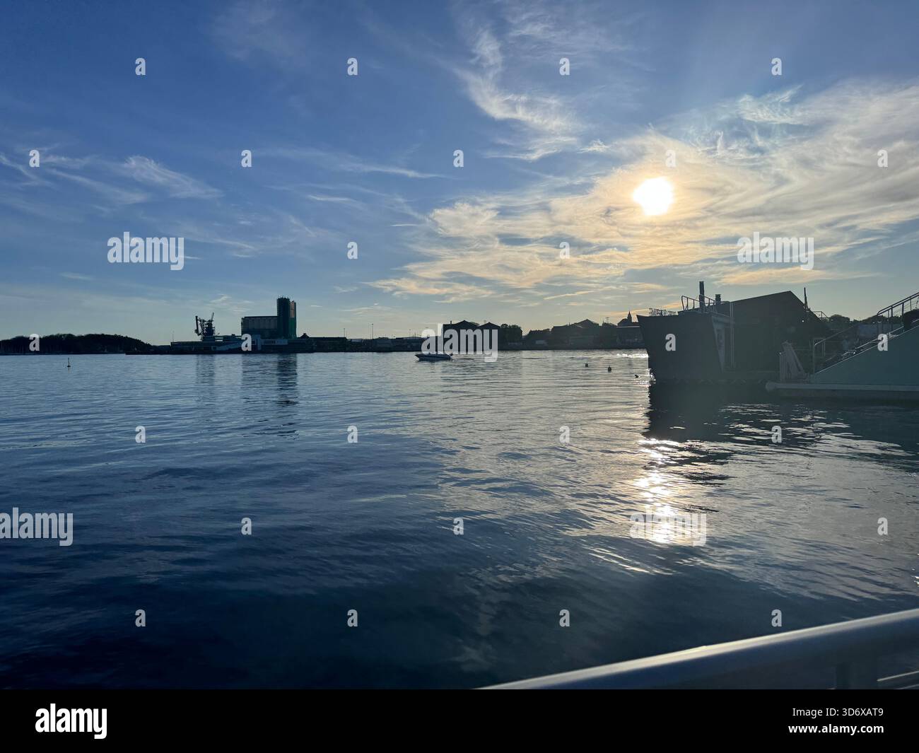 La douce lumière dorée du soleil se reflète sur l'eau calme tandis que le soleil se couche derrière des bâtiments lointains et des nuages dérivants. - Image de stock capturée avec un smartphone