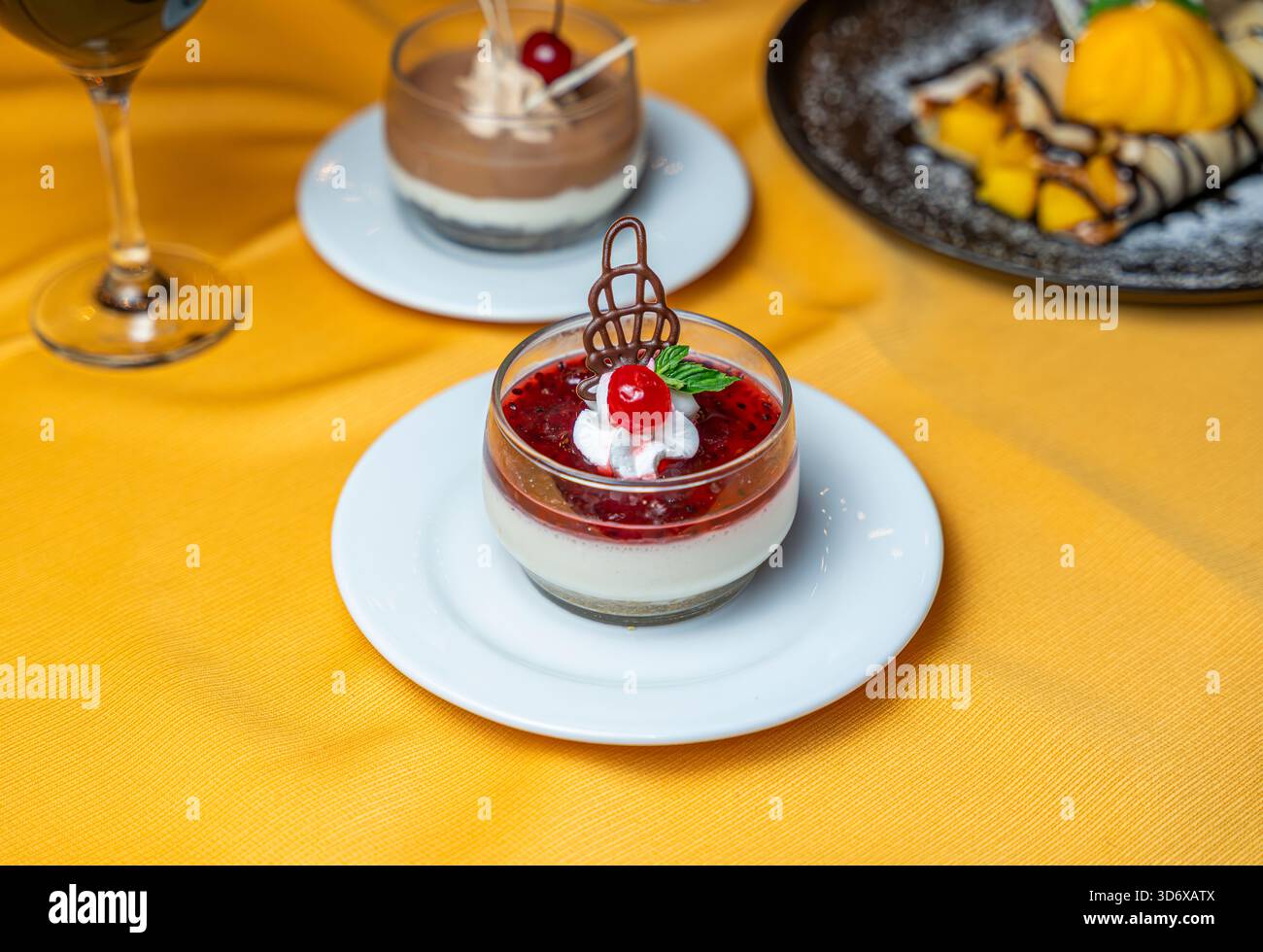 Dessert à la panna cotta aux baies garni de cerise, crème fouettée, menthe et décoration chocolat servi dans une tasse en verre sur une assiette blanche sur un tissu jaune. Banque D'Images
