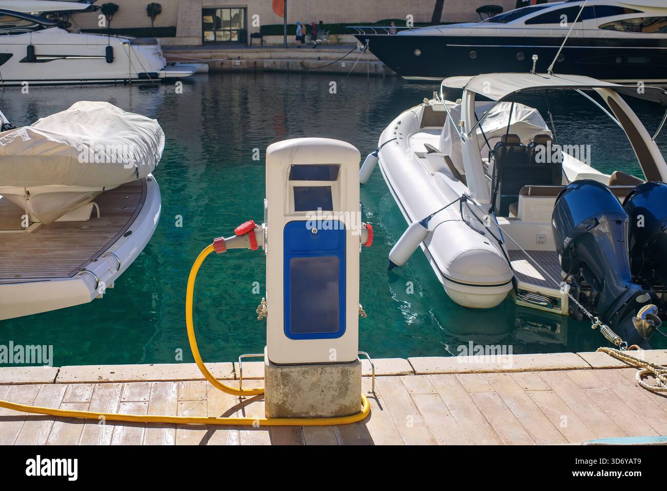 Station de ravitaillement avec un long tuyau jaune sur un quai en bois d'une marina de luxe, avec des yachts amarrés et des bateaux dans une eau turquoise brillante. Banque D'Images