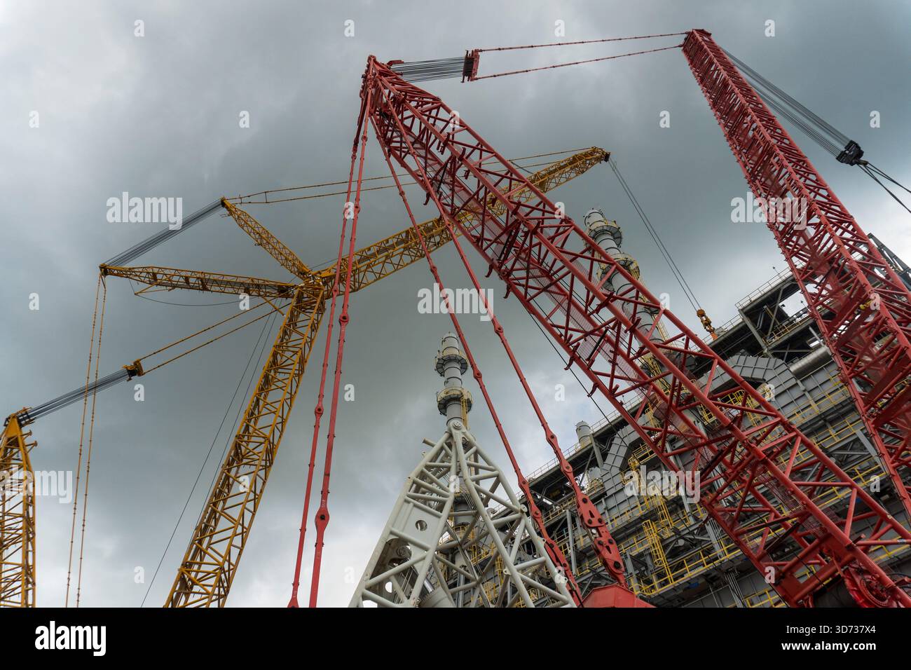 Grue sur chenilles massive avec des flèches en treillis rouge positionnées sur un site de construction de raffinerie, soulevant de l'équipement près de hautes tours de traitement sous un ski nuageux Banque D'Images